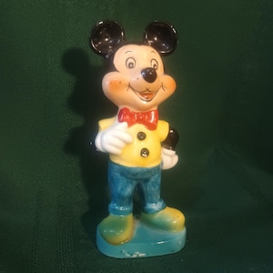 May include: A ceramic Mickey Mouse figurine. The figure is wearing a yellow shirt, blue pants, and green shoes. It has a red bow tie and black ears. The figurine is standing on a blue base.