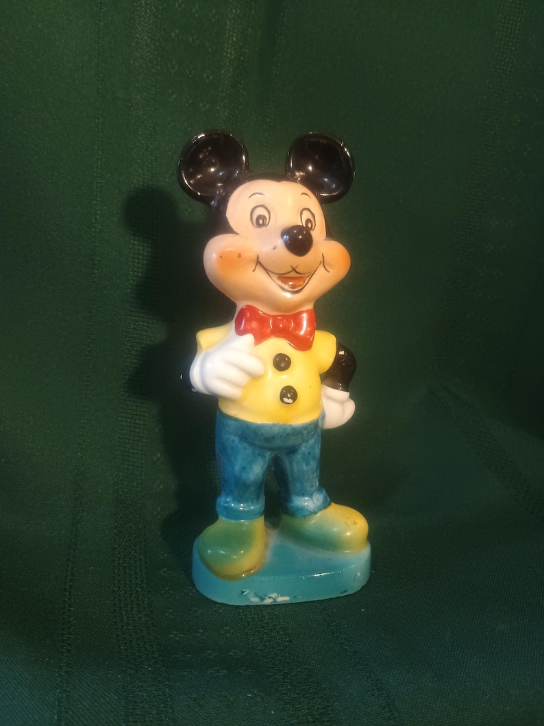 May include: A ceramic Mickey Mouse figurine. The figure is wearing a yellow shirt, blue pants, and green shoes. It has a red bow tie and black ears. The figurine is standing on a blue base.