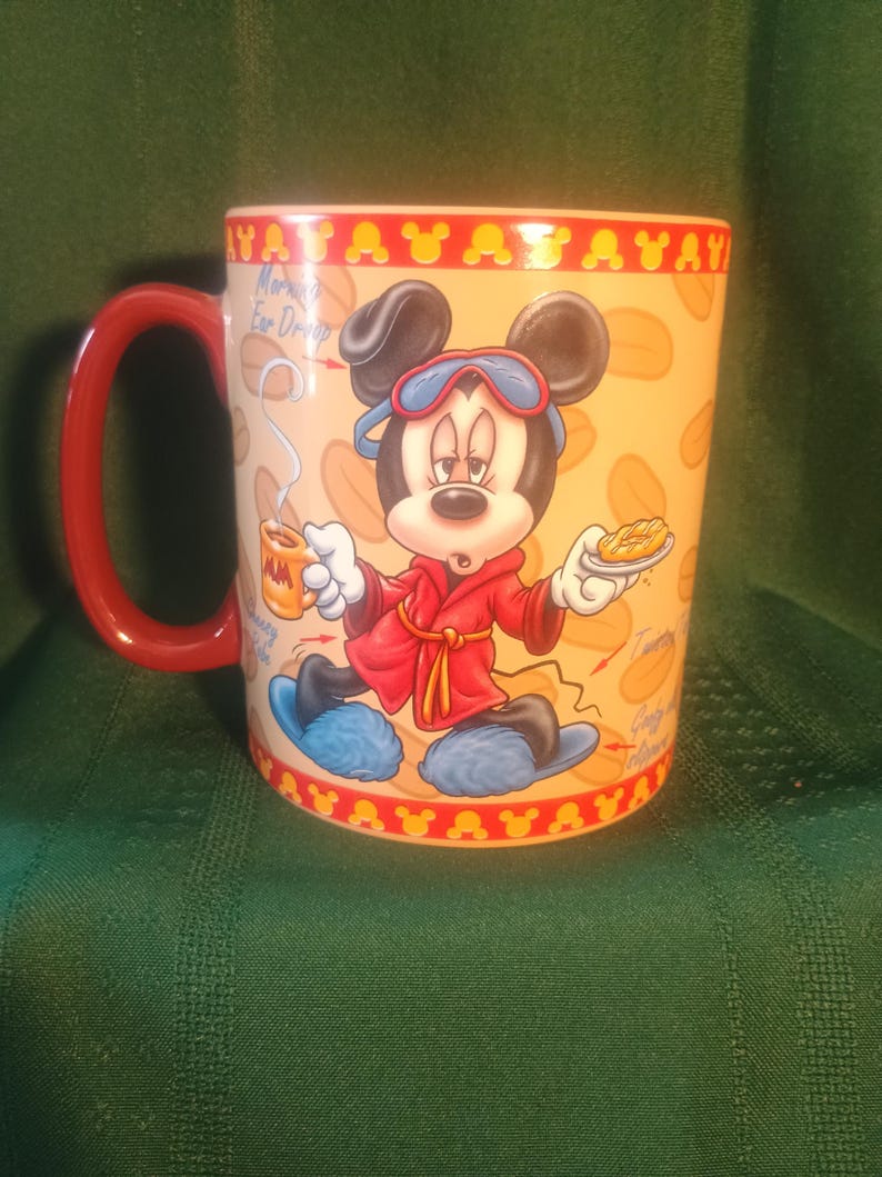 Może przedstawiać: Ceramiczny kubek do kawy z Myszka Miki w czerwonym szlafroku i niebieskich goglach. Kubek ma czerwony uchwyt i ż&oacute;łte wnętrze. Projekt zawiera tekst "Morning Ear Drops".