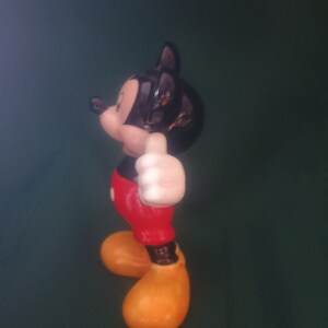 Puede incluir: Una figura de cer&aacute;mica de Mickey Mouse, un personaje cl&aacute;sico de Disney. Mickey est&aacute; representado con su atuendo ic&oacute;nico: pantalones cortos rojos, zapatos amarillos, guantes blancos y cabeza negra. Est&aacute; haciendo un gesto de pulgar hacia arriba.