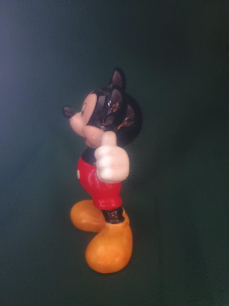 Puede incluir: Una figura de cer&aacute;mica de Mickey Mouse, un personaje cl&aacute;sico de Disney. Mickey est&aacute; representado con su atuendo ic&oacute;nico: pantalones cortos rojos, zapatos amarillos, guantes blancos y cabeza negra. Est&aacute; haciendo un gesto de pulgar hacia arriba.