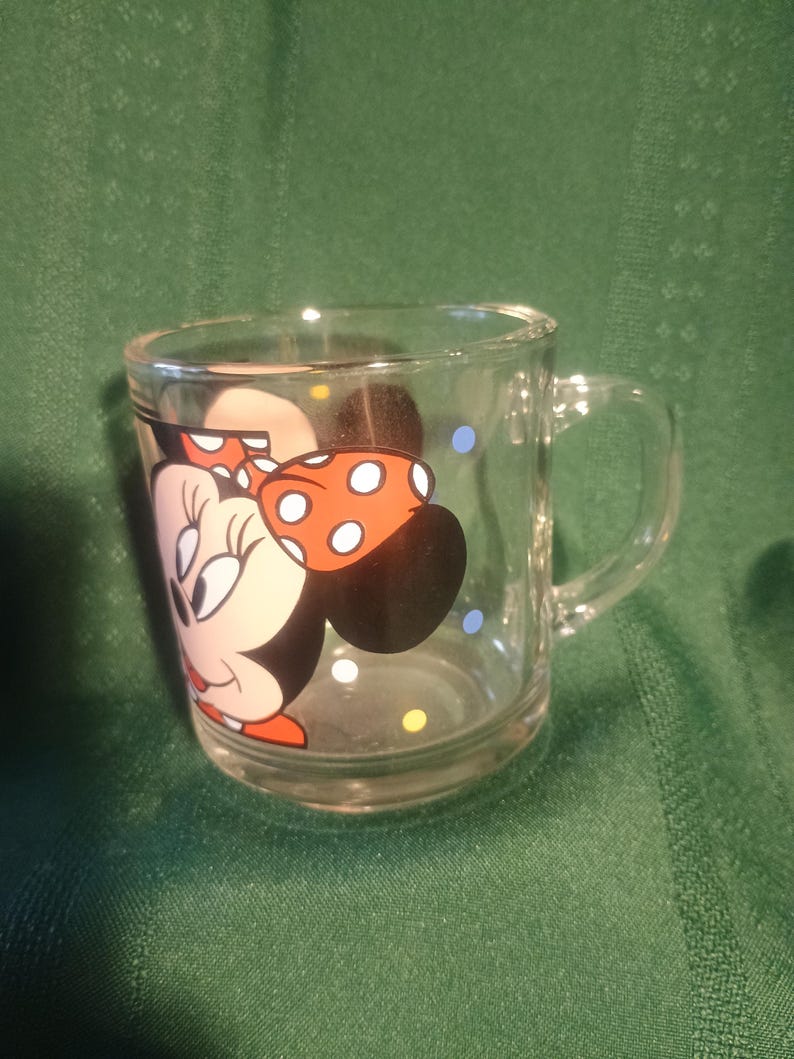 Pu&ograve; includere: Tazza di vetro trasparente con un'immagine a cartoni animati di Minnie Mouse. Il design include il volto di Minnie, un fiocco rosso a pois bianchi e pois colorati. La tazza ha un manico ed &egrave; adatta alle bevande.