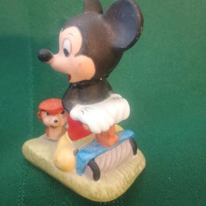 Op de afbeelding: Een keramieken beeldje van Mickey Mouse in een klassieke pose, met een rode broek, gele schoenen en een witte tutu. Een klein beeldje van Pluto is ook aanwezig. De basis is lichtgeel van kleur en de achtergrond is groen.