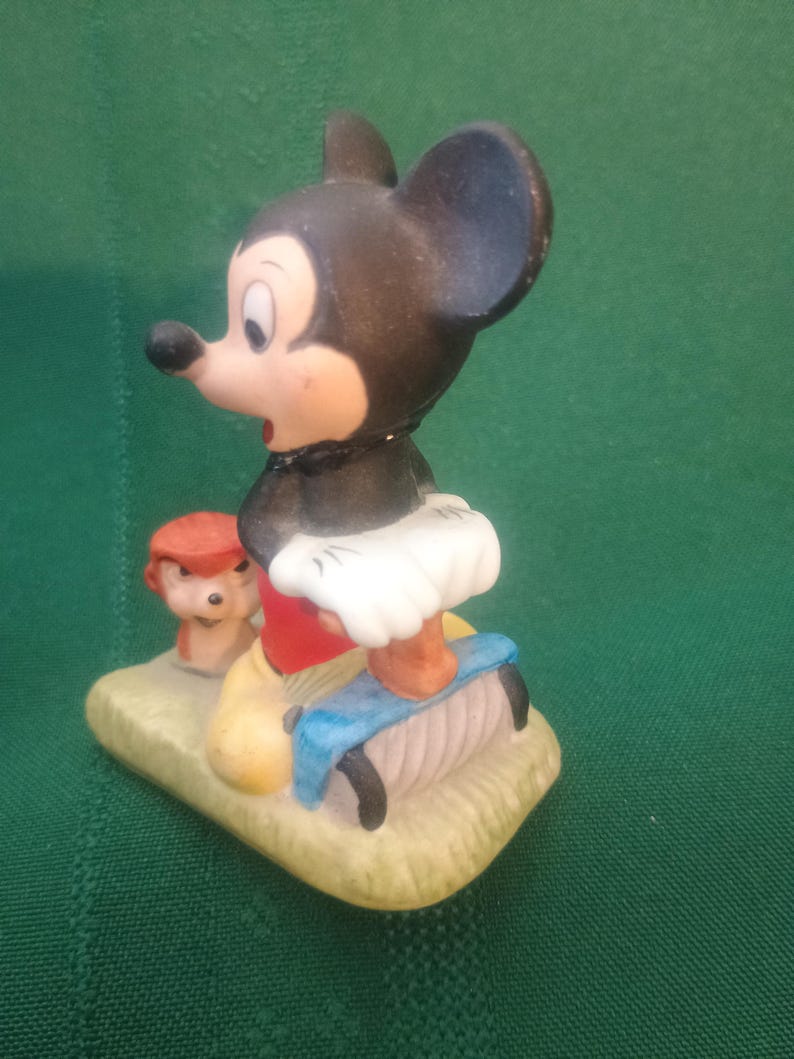 Op de afbeelding: Een keramieken beeldje van Mickey Mouse in een klassieke pose, met een rode broek, gele schoenen en een witte tutu. Een klein beeldje van Pluto is ook aanwezig. De basis is lichtgeel van kleur en de achtergrond is groen.
