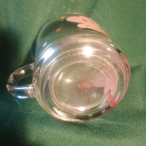 Pu&ograve; includere: Tazza di vetro trasparente con manico, con un design bianco e rosa con punti rossi e gialli. La tazza &egrave; posta su uno sfondo verde, evidenziando la sua trasparenza e il design.