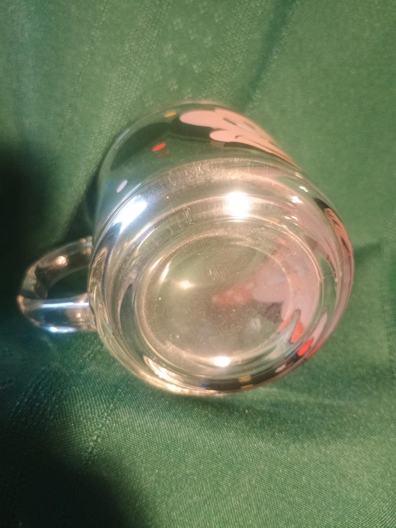 Pu&ograve; includere: Tazza di vetro trasparente con manico, con un design bianco e rosa con punti rossi e gialli. La tazza &egrave; posta su uno sfondo verde, evidenziando la sua trasparenza e il design.