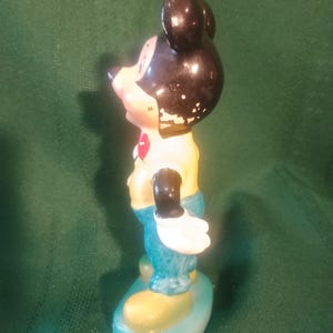 May include: Vintage Mickey Mouse figurine. The figure has a black head and ears, a yellow shirt, blue pants, and white gloves. The figurine is standing on a blue and yellow base. The paint shows signs of wear.