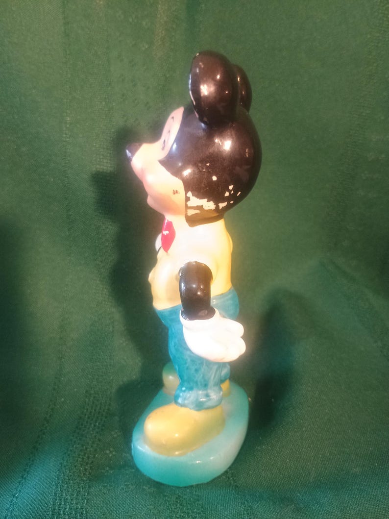 May include: Vintage Mickey Mouse figurine. The figure has a black head and ears, a yellow shirt, blue pants, and white gloves. The figurine is standing on a blue and yellow base. The paint shows signs of wear.