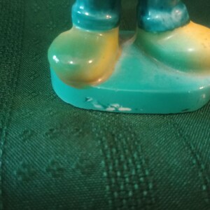 May include: A ceramic figurine featuring teal pants and yellow and green shoes. The figurine is standing on a teal base. The paint has some wear and tear.