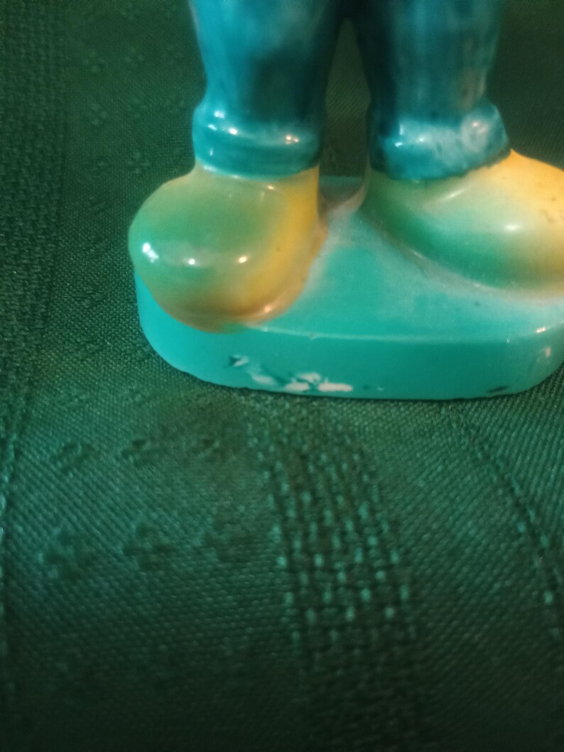 May include: A ceramic figurine featuring teal pants and yellow and green shoes. The figurine is standing on a teal base. The paint has some wear and tear.