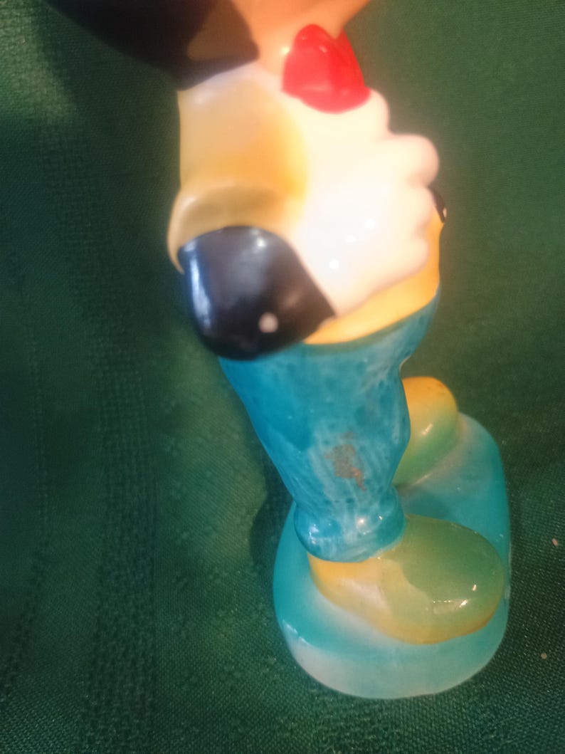 May include: A vintage figurine of a cartoon character, featuring a red bow tie, yellow shirt, and blue pants. The figurine has a white face and black ears. The base and shoes are light blue and yellow.