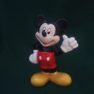 Puede incluir: Una figura de cer&aacute;mica de Mickey Mouse, un personaje cl&aacute;sico de Disney. Mickey se representa con orejas negras, cuerpo negro, pantalones cortos rojos con botones blancos, guantes blancos y zapatos amarillos. Est&aacute; haciendo una se&ntilde;al de pulgar.