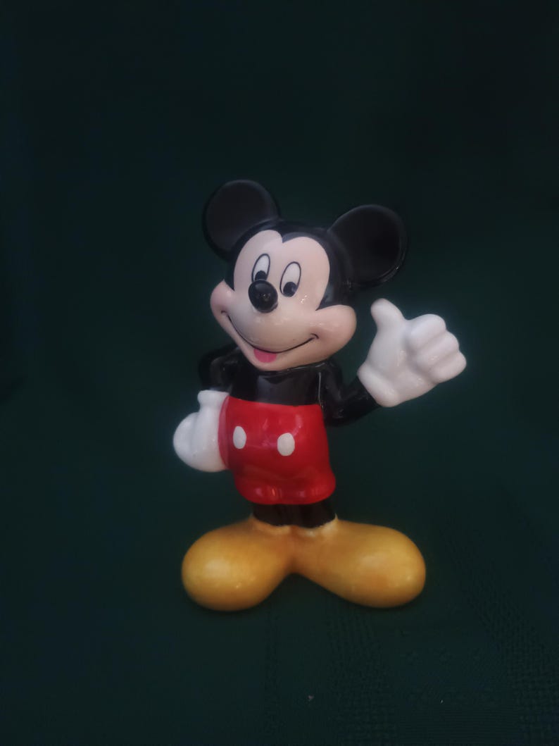 Puede incluir: Una figura de cer&aacute;mica de Mickey Mouse, un personaje cl&aacute;sico de Disney. Mickey se representa con orejas negras, cuerpo negro, pantalones cortos rojos con botones blancos, guantes blancos y zapatos amarillos. Est&aacute; haciendo una se&ntilde;al de pulgar.
