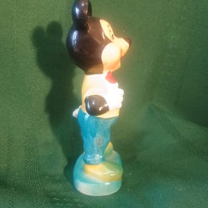 May include: A ceramic figurine of Mickey Mouse, featuring a black head and ears, a tan face, and a red bow tie. He wears a white shirt, black gloves, blue pants, and yellow shoes. The figurine is standing on a light blue base.