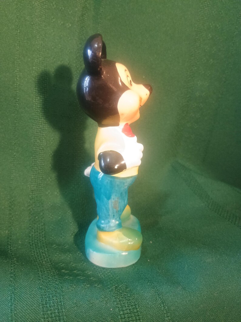 May include: A ceramic figurine of Mickey Mouse, featuring a black head and ears, a tan face, and a red bow tie. He wears a white shirt, black gloves, blue pants, and yellow shoes. The figurine is standing on a light blue base.
