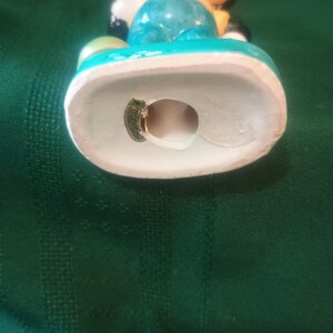 May include: A vintage ceramic figurine of a cartoon character, featuring a light blue body, yellow shirt, and black shoes. The figurine has a white base with a turquoise trim. The character's face is peach-colored with black eyes and a red mouth.