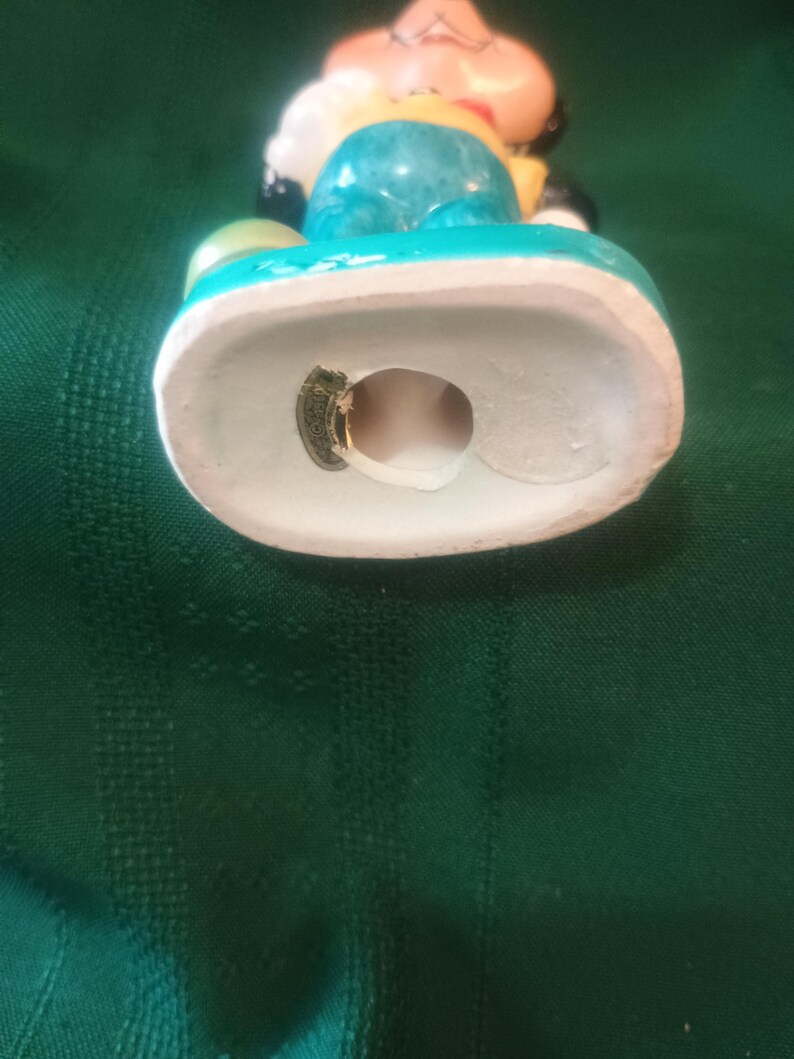 May include: A vintage ceramic figurine of a cartoon character, featuring a light blue body, yellow shirt, and black shoes. The figurine has a white base with a turquoise trim. The character's face is peach-colored with black eyes and a red mouth.