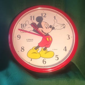 May include: A red-rimmed Lorus Quartz clock featuring Mickey Mouse. The clock face is white with black numerals and red hands. Mickey Mouse is depicted in his classic outfit, with yellow shoes, red shorts, and black gloves.