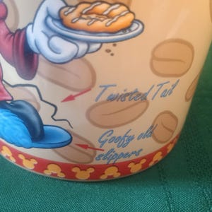 Może przedstawiać: Beżowy ceramiczny kubek z postacią z kresk&oacute;wki trzymającą pączka. Kubek ma napis "Twisted Tail" i "Goofy old slippers". Podstawa kubka ma czerwony brzeg z ż&oacute;łtymi sylwetkami Myszki Miki.
