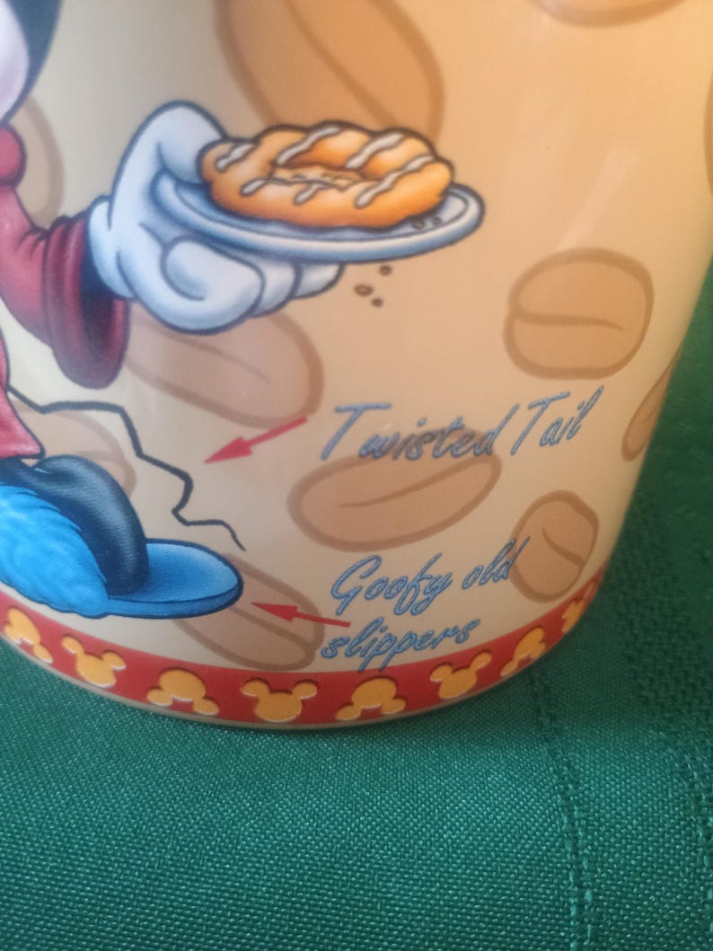 Może przedstawiać: Beżowy ceramiczny kubek z postacią z kresk&oacute;wki trzymającą pączka. Kubek ma napis "Twisted Tail" i "Goofy old slippers". Podstawa kubka ma czerwony brzeg z ż&oacute;łtymi sylwetkami Myszki Miki.