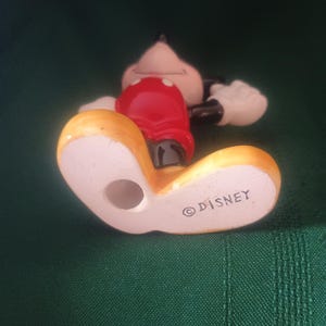 Puede incluir: Una figura de cer&aacute;mica de Mickey Mouse, tumbada, con una camisa roja, pantalones cortos negros y zapatos amarillos. La figura tiene guantes blancos y una cola negra. La palabra "DISNEY" est&aacute; impresa en la parte inferior del zapato.