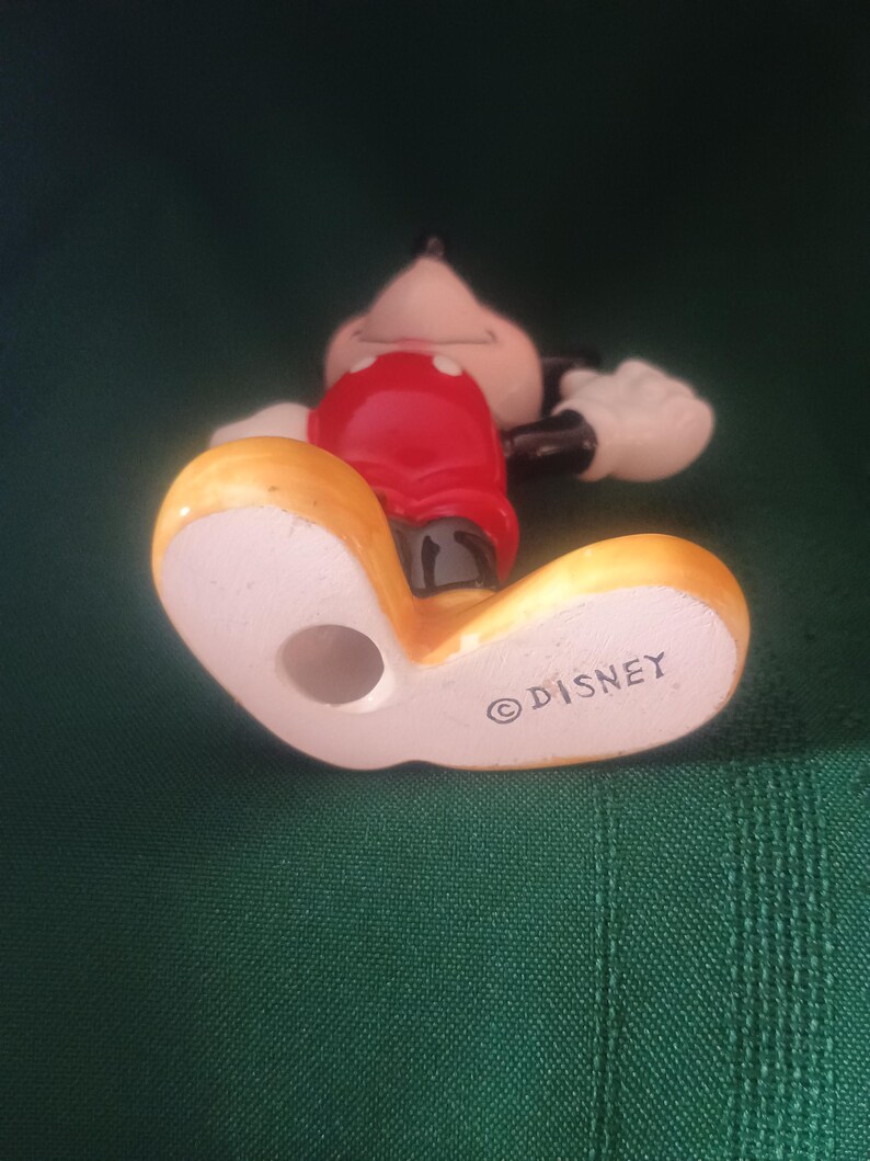 Puede incluir: Una figura de cer&aacute;mica de Mickey Mouse, tumbada, con una camisa roja, pantalones cortos negros y zapatos amarillos. La figura tiene guantes blancos y una cola negra. La palabra "DISNEY" est&aacute; impresa en la parte inferior del zapato.