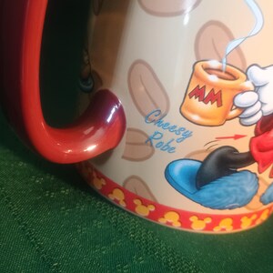 Może przedstawiać: Ceramiczny kubek do kawy z czerwonym uchwytem i obramowaniem, z motywem postaci z kresk&oacute;wek. Kubek ma beżowe tło z brązowymi kształtami i postać z kresk&oacute;wki trzymającą mały pomarańczowy kubek. Na kubku widnieje napis "Cheesy Robe".