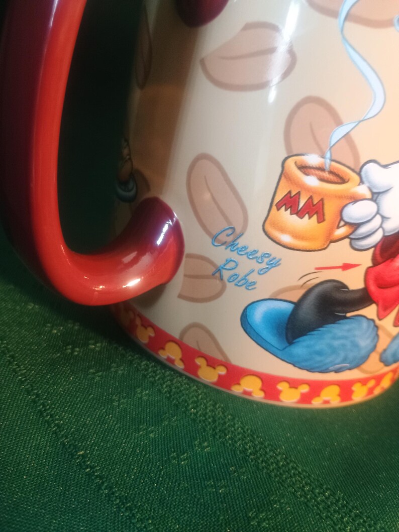 Może przedstawiać: Ceramiczny kubek do kawy z czerwonym uchwytem i obramowaniem, z motywem postaci z kresk&oacute;wek. Kubek ma beżowe tło z brązowymi kształtami i postać z kresk&oacute;wki trzymającą mały pomarańczowy kubek. Na kubku widnieje napis "Cheesy Robe".