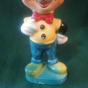 May include: A vintage Mickey Mouse figurine. The ceramic figure features a yellow shirt with black buttons, blue pants, and green shoes. Mickey has a red bow tie and white gloves. The figurine is standing on a blue base.