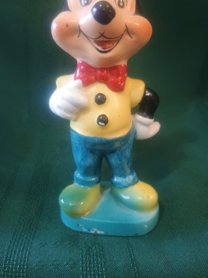 May include: A vintage Mickey Mouse figurine. The ceramic figure features a yellow shirt with black buttons, blue pants, and green shoes. Mickey has a red bow tie and white gloves. The figurine is standing on a blue base.