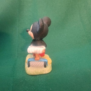 Op de afbeelding: Een klein beeldje van een tekenfilmmuis met zwarte oren en een zwart lichaam. De muis draagt een witte rok en rode schoenen. Hij staat op een gele basis met een blauw-witte bank.