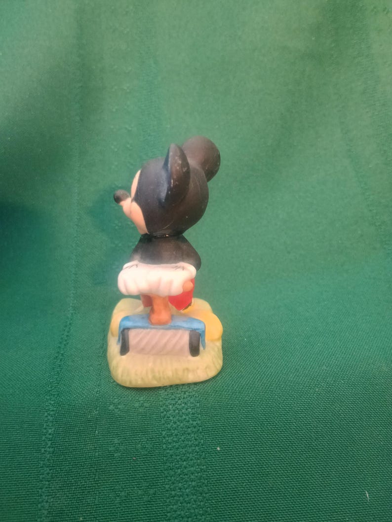 Op de afbeelding: Een klein beeldje van een tekenfilmmuis met zwarte oren en een zwart lichaam. De muis draagt een witte rok en rode schoenen. Hij staat op een gele basis met een blauw-witte bank.