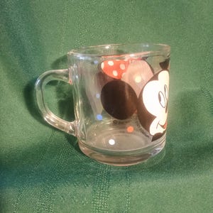 Pu&ograve; includere: Tazza di vetro trasparente con un disegno di Topolino. La tazza ha un manico e una base arrotondata. Topolino &egrave; raffigurato con un viso nero, guanti bianchi e un fiocco a pois rossi e bianchi. La tazza &egrave; di The Walt Disney Company.