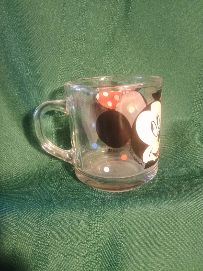 Pu&ograve; includere: Tazza di vetro trasparente con un disegno di Topolino. La tazza ha un manico e una base arrotondata. Topolino &egrave; raffigurato con un viso nero, guanti bianchi e un fiocco a pois rossi e bianchi. La tazza &egrave; di The Walt Disney Company.