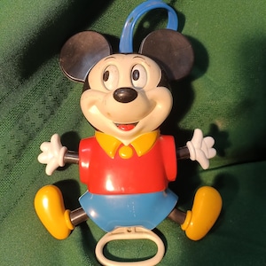 May include: A plastic Mickey Mouse toy featuring a black head with large round ears, a red shirt with a yellow collar, and blue shorts. The figure has white gloves, yellow shoes, a blue hanging loop at the top, and a white pull-ring at the bottom.