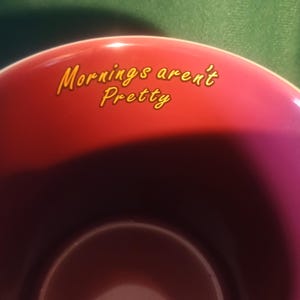 Może przedstawiać: Bordowa ceramiczna filiżanka z napisem "Mornings aren't Pretty" w ż&oacute;łtym skrypcie. Filiżanka ma ciemne wnętrze i błyszczące wykończenie, idealna do kawy lub herbaty.