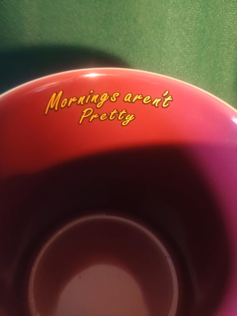 Może przedstawiać: Bordowa ceramiczna filiżanka z napisem "Mornings aren't Pretty" w ż&oacute;łtym skrypcie. Filiżanka ma ciemne wnętrze i błyszczące wykończenie, idealna do kawy lub herbaty.