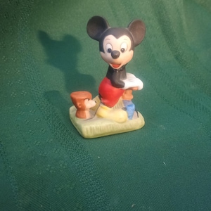 Op de afbeelding: Een keramieken beeldje van Mickey Mouse in een klassieke pose, gekleed in een rode broek, zwarte jas en gele schoenen. Een kleine bruine hondenfiguur staat naast hem. De figuren staan op een groene basis.