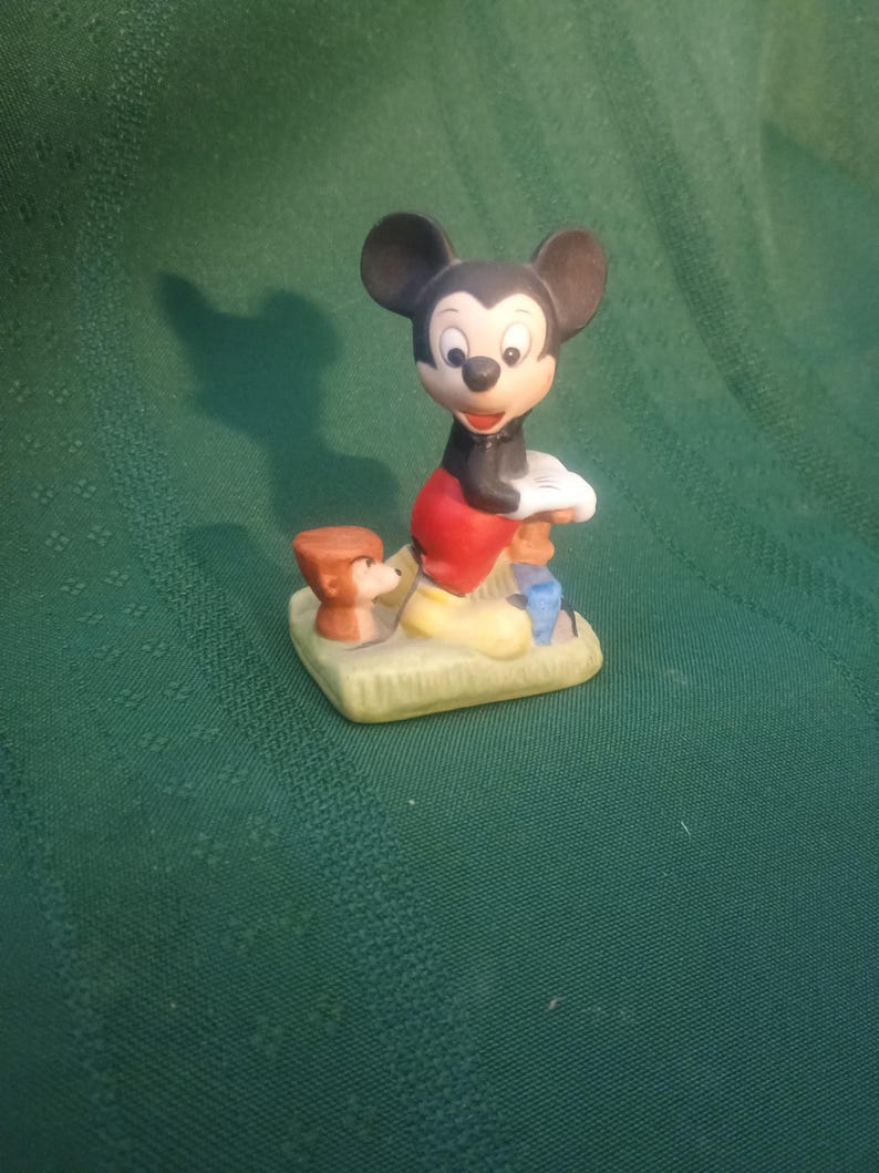 Op de afbeelding: Een keramieken beeldje van Mickey Mouse in een klassieke pose, gekleed in een rode broek, zwarte jas en gele schoenen. Een kleine bruine hondenfiguur staat naast hem. De figuren staan op een groene basis.
