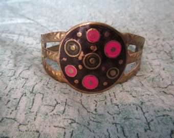 May include: A gold-toned bracelet with a circular design featuring pink and black circles. The bracelet has a wide band with three open spaces.