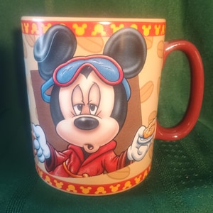 Może przedstawiać: Ceramiczny kubek do kawy z Myszka Miki w czerwonej kurtce i niebieskich goglach. Kubek ma czerwony uchwyt i beżowe tło z powtarzającym się wzorem gł&oacute;w Myszki Miki i słowem "YA".