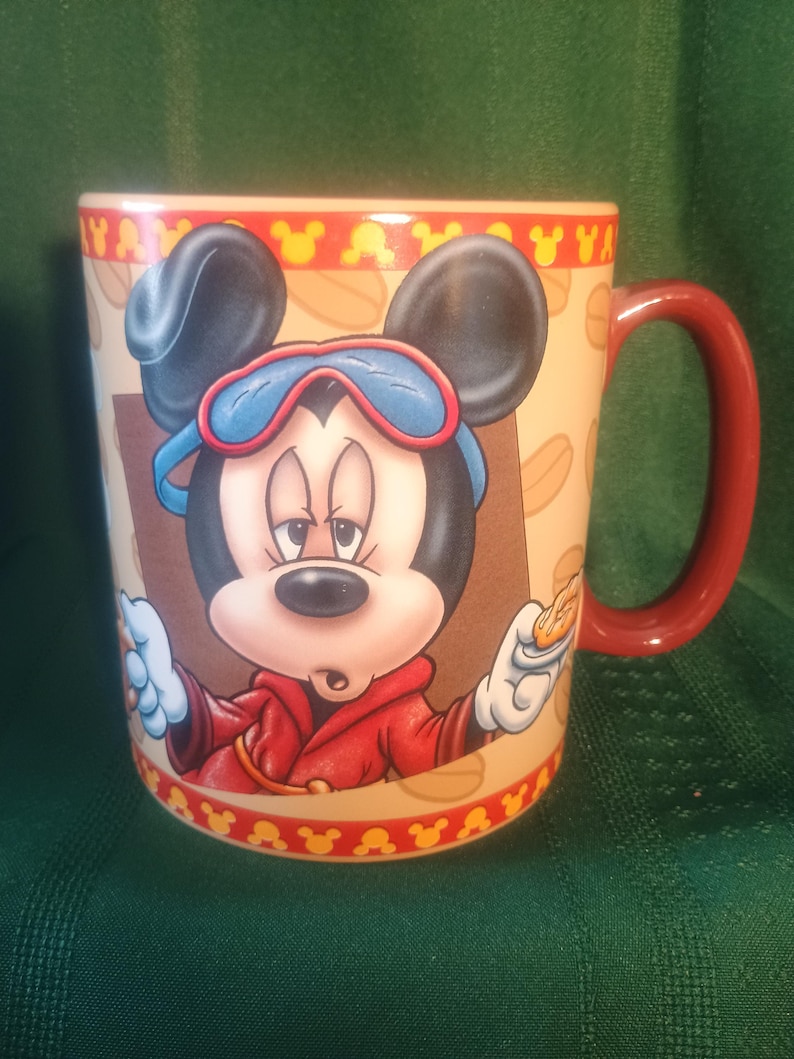 Może przedstawiać: Ceramiczny kubek do kawy z Myszka Miki w czerwonej kurtce i niebieskich goglach. Kubek ma czerwony uchwyt i beżowe tło z powtarzającym się wzorem gł&oacute;w Myszki Miki i słowem "YA".