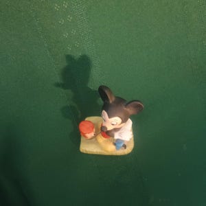Op de afbeelding: Een kleine, keramische Mickey Mouse-beeldje. Mickey is afgebeeld in zijn klassieke outfit, met zwarte oren, een wit overhemd, gele schoenen en een rode broek. Hij houdt een klein, oranje voorwerp vast. Het beeldje staat op een kleine, gele basis.
