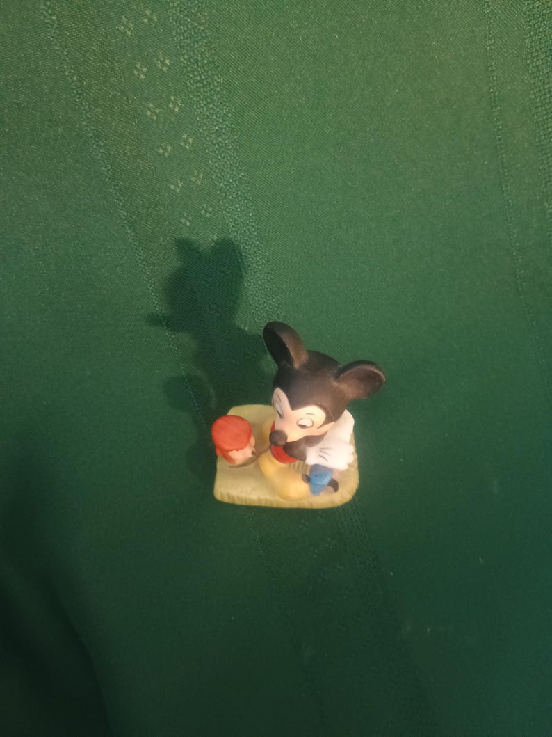 Op de afbeelding: Een kleine, keramische Mickey Mouse-beeldje. Mickey is afgebeeld in zijn klassieke outfit, met zwarte oren, een wit overhemd, gele schoenen en een rode broek. Hij houdt een klein, oranje voorwerp vast. Het beeldje staat op een kleine, gele basis.