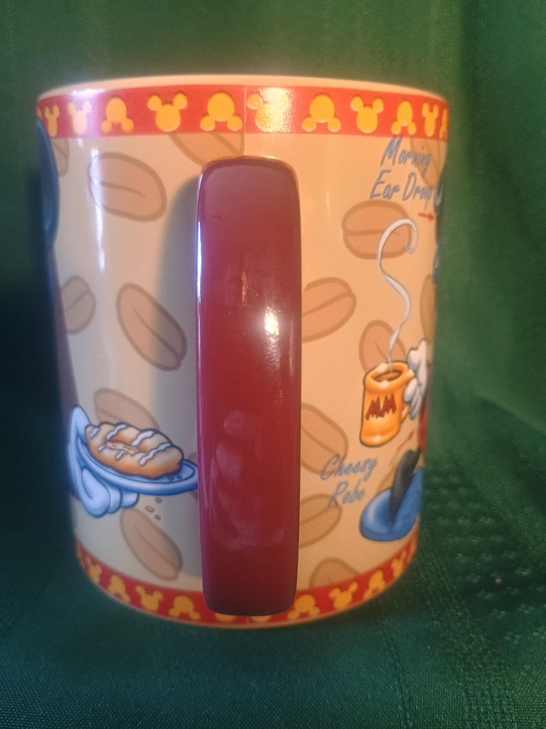 Może przedstawiać: Ceramiczny kubek do kawy z czerwonym uchwytem. Kubek ma beżowe tło z ilustracjami ziaren kawy i czerwone obramowanie z sylwetkami Myszki Miki. Kubek ma tekst i ilustracje Myszki Miki.