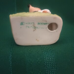 Op de afbeelding: Vintage keramieken beeldje met een personage met een bruin gezicht, zwart haar en een witte hoed, op een groene basis. De basis heeft de tekst "&copy; WALT DISNEY PRODUCTIONS". Het beeldje staat tegen een groene achtergrond.