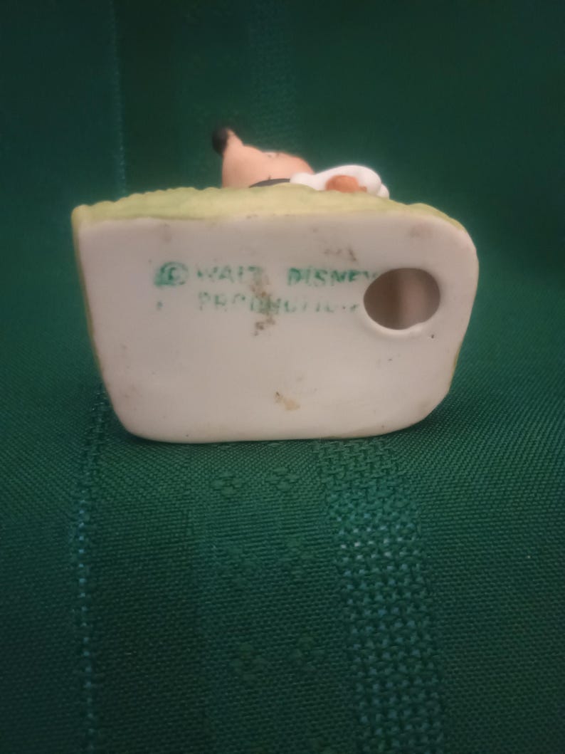 Op de afbeelding: Vintage keramieken beeldje met een personage met een bruin gezicht, zwart haar en een witte hoed, op een groene basis. De basis heeft de tekst "&copy; WALT DISNEY PRODUCTIONS". Het beeldje staat tegen een groene achtergrond.