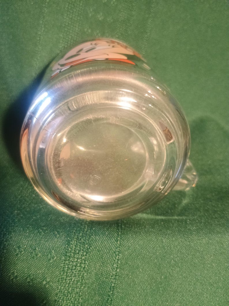 Pu&ograve; includere: Tazza di vetro trasparente con un piccolo manico. La tazza ha un disegno sul bordo superiore con colori arancione, bianco e beige. La tazza &egrave; appoggiata su una superficie verde.