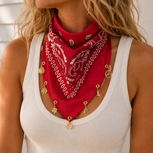 May include: A red bandana featuring a white paisley pattern, adorned with several small gold-toned charms hanging from the bottom edge. The charms include a sun, a mountain, a boot, a four-leaf clover, a pistol, and a horseshoe, all attached to the fabric.