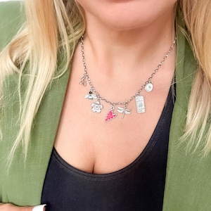 May include: A silver-toned charm necklace featuring multiple dangling pendants including a bee, a flower, a pink heart, a dragonfly, and a rectangular tag with celestial symbols. The chain is a standard link style with a secure clasp mechanism.